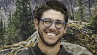 Headshot of a man with dark hair and glasses standing in front of a forest background