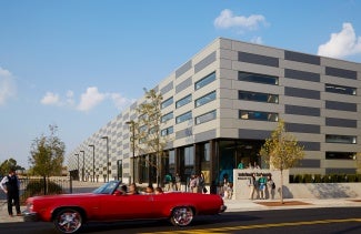 School exterior At day. The school has an alternating color facade of Gray and beige. There is a red car in the foreground. 