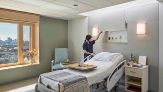 A nurse in dark scrubs adjusts a shade in a patient room. 