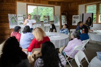 A woman gives a presentation to a large group of people sitting at round tables. 