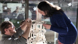 A man and a woman construct a tall model together in front of a glass wall.