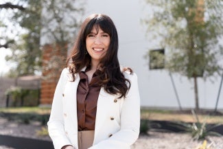 Headshot of a woman with long dark hair in a white blazer standing outside a building.