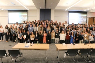 A large group of people poses together in a meeting room with projection screens in the background.