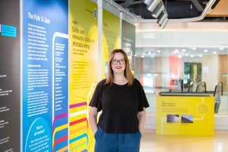 Woman with long hair and glasses in a black t-shirt stands in front on an exhibit.