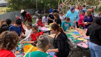 A large group of children work on art projects outside at various tables. There is a playground in the background.