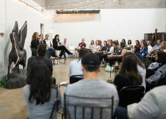 A panel of four women give a presentation in front of an audience in a white-walled gallery space next to a Pegasus statue.