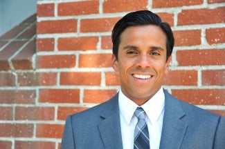 Headshot of a man with dark hair, wearing a gray suit and standing in front of a brick wall. 