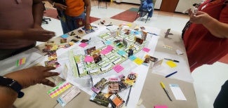 Photo of a table with an architectural plan on it. The plan is covered in sticky notes. You can see the hands of four people standing around the table out of frame. 