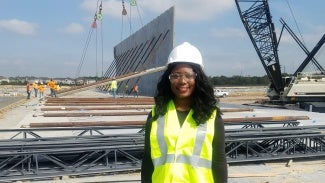 A woman in a visibility vest and a hard hat stands on a construction site. 