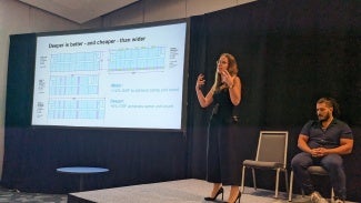 A woman is giving a presentation on stage at an event space next to a projector screen. 