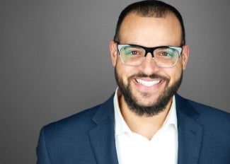 Headshot of a man wearing glasses and a navy blazer standing in front of a Gray background. 