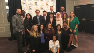 A group of people pose in front of a step and repeat backdrop in a banquet room. 