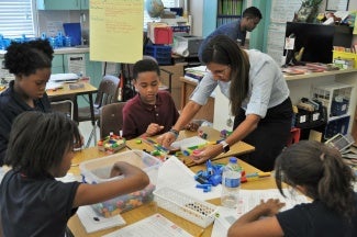 Four children sit around a classroom table with an instructor in the center. The instructor is helping them with an art project.