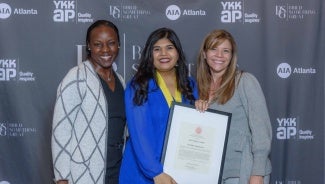 Three women in front of a step and repeat backdrop at an event. The woman in the center is wearing a medal and holding a framed certificate.