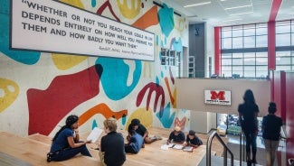 Students sit on bleacher steps next to a wall mural with an inspirational quote.