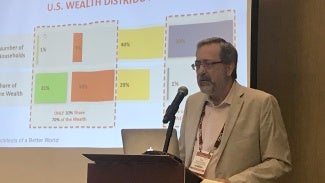 A man with glasses and a beard presents at a lectern with a chart projected behind him.