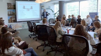 View of a classroom from the back. There is a boardroom table in the middle of the room with rolling chairs. Students sit at the table as well as along the sides of the room. A teacher is standing in the front of the classroom next to a projection screen giving a lecture.