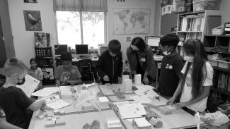 A group of children work with an instructor around a table in a classroom. There are building models on the table.