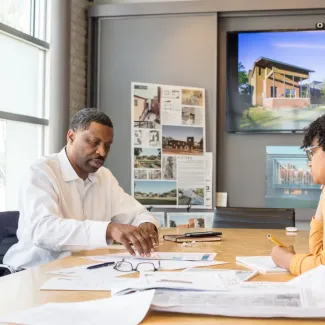 Three employees sit around a conference table with papers laid in front of them, on the screen behind them is an image of a building