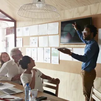 One employee gives a presentation at a board while other employees watch seated at a conference table