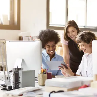 Three young employees smile as they all look over paperwork together at a desk in a sunny office