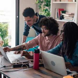 Three smiling employees sit and stand in front of a desk with two laptops open, behind them is a bookshelf