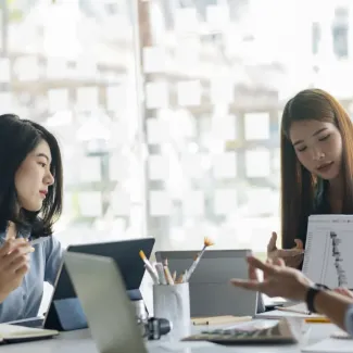 Three employees sit around the end of a conference table with laptops and paper, behind them is a brightly lit window