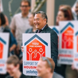 A group of people stand holding signs in favor of affordable housing. Douglas Ito stands center.