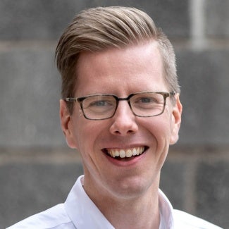 headshot of a man with short blonde hair wearing glasses and a white shirt.