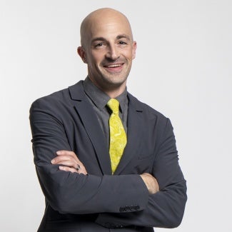 Headshot of a bald man in a dark jacket with a yellow tie standing in front of a gray background