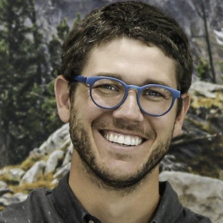 Headshot of a man with dark hair and glasses standing in front of a forest background