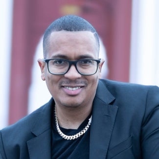 Headshot of a man with short hair in a black blazer wearing a chain necklace and glasses. A red bridge is visible in the background.