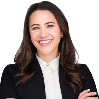 A woman with long dark hair in a black blazer stands in front of a white background.