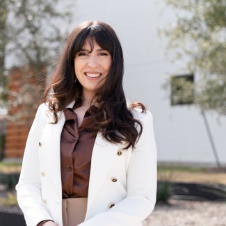 Headshot of a woman with long dark hair in a white blazer standing outside a building.
