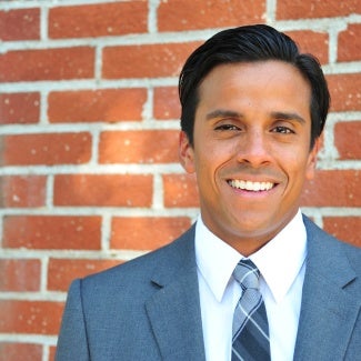 Headshot of a man with dark hair, wearing a gray suit and standing in front of a brick wall. 