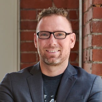 Headshot of a man wearing glasses and a blazer over a t-shirt.