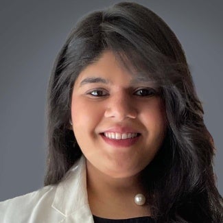 headshot of a woman with long dark hair and a white blazer in front of a grey background.