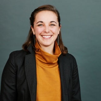 Headshot of a woman with long brown hair, wearing an orange sweater and a dark blazer. 