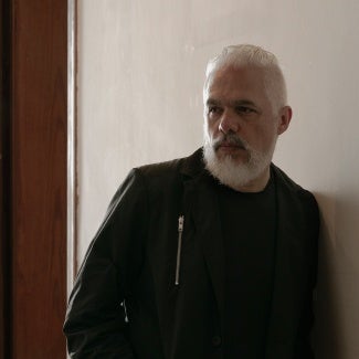 man with white hair an beard in a black leather jacket standing in front of a light colored wall.