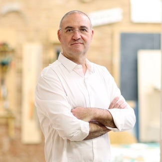 man in wireframe glasses in a light colored button down shirt stands in front of a sandstone building.