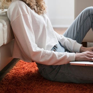 Woman siting on the floor against a couch with a laptop computer in her lap.