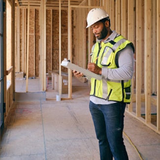 Man wearing PPE stands in wooden-framed construction site, looking at his clipboard