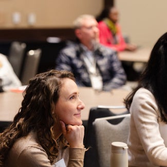 Participants sitting at table, listening intently to speaker. Two women in main focus, with other people blurred in the foreground and background.