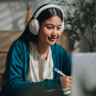Lady sitting at a computer studying for exam, while taking notes and listening to headphones